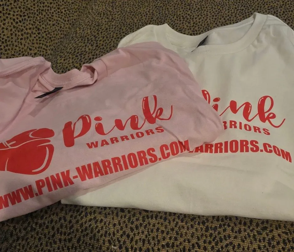Two folded Pink Warriors T-shirts, one pink and one white, printed in red with the Pink Warriors logo and www.pink-warriors.com